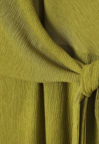 Tissu vert olive texturé noué en un nœud, montrant des motifs côtelés et froissés détaillés.