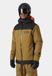 Veste de ski en marron et noir, avec une fermeture éclair à l'avant, un col haut, des poignets élastiques et un logo sur la poitrine. Associée à des gants noirs.