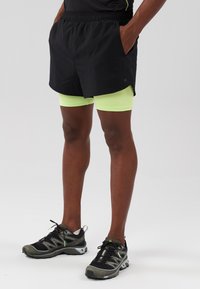 Pantaloni da ginnastica neri con cintura elastica, tasche laterali e pantaloni interni verde neon. Abbinati a scarpe da corsa nere e grigie.