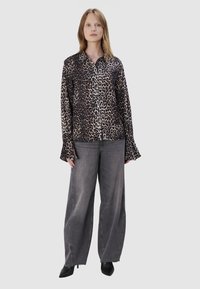 Blouse met luipaardprint en lange mouwen, met een knoopsluiting aan de voorkant, gecombineerd met grijze wijde jeans en zwarte enkellaarsjes. Effen achtergrond.