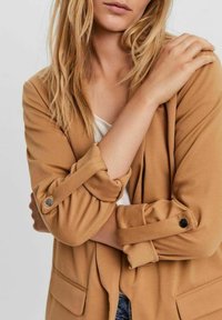 Dlouhý blazer s dlouhým rukávem z měkké látky s ohrnutými manžetami a zapínáním na druky. Má dvě přední kapsy a volný střih.