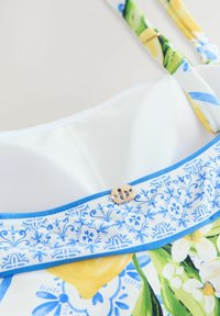 Haut de bikini à motifs floraux en blanc et bleu, avec des accents jaunes, des bretelles ajustables et un fermoir doré à l'avant.