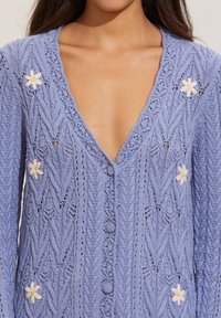 Světle fialový pletený svetr se zapínáním na knoflíky do V, s texturovaným vzorem, zelenými knoflíky a květinovými ozdobami u výstřihu a kapes.
