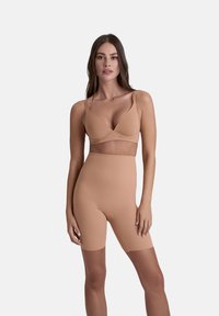 Donna che indossa una shapewear color carne, tra cui un perizoma lungo a vita alta e un reggiseno abbinato, in piedi contro uno sfondo neutro.