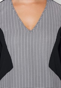 Černobílý top ve stylu V-neck s houndstooth vzorem na přední straně a jednobarevnými černými rukávy, s hladkou texturou a čistými liniemi.