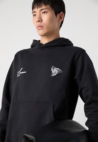 Hoodie noir en tissu doux, avec un logo brodé blanc et un design géométrique sur la poitrine. Comprend une poche avant.