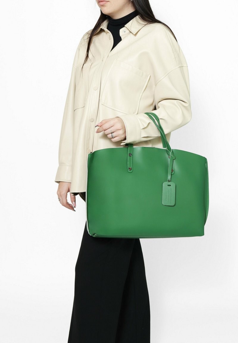Sac tote en cuir vert avec double anses, texture lisse, forme rectangulaire et une étiquette attachée. Porté avec une chemise beige et un pantalon noir.