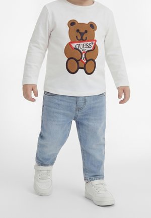 Småbarn iført hvid langærmet skjorte med brun bamse, der holder en "GUESS" undertøjsgrafik, lyseblå jeans og hvide sneakers.
