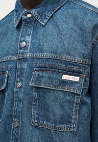 Εγγύς λήψη ενός ατόμου που φοράει μπλε τζιν μπουφάν με τσέπες με καπάκι και μικρή ετικέτα Calvin Klein στην αριστερή τσέπη στο στήθος.