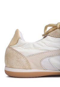 Beige and white athletic shoe featuring a suede and leather blend, textured accents, flat laces, and a rubber sole. Rounded design at the heel.