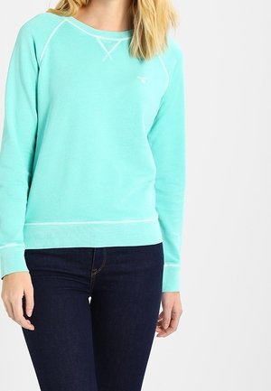 Frau trägt ein helltürkisfarbenes Sweatshirt mit langen Ärmeln und weißen Nähten, kombiniert mit dunkelblauen Jeans vor einem einfarbigen weißen Hintergrund.