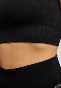 Crop top noir à côtes avec un design ajusté, présentant une large bande en bas et une texture lisse. La coupe inclut une exposition du ventre.