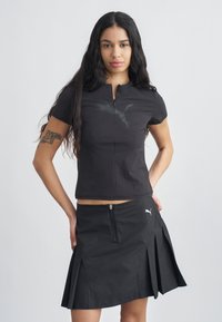 Νέα γυναίκα με μακριά σκούρα μαλλιά, που φοράει μαύρο φερμουάρ Puma και πλισέ φούστα, στέκεται με τα χέρια στη μέση μπροστά σε απλό φόντο.