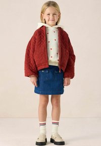 Rode gewatteerde bomberjack met een rits, witte hoodie met rode hartpatronen, spijkerrok, gestreepte sokken en beige loafer schoenen.