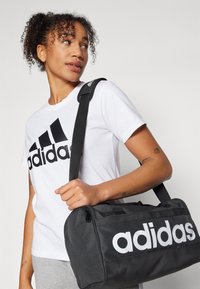Svart duffelbag med hvit "adidas"-logo, laget av slitesterkt materiale. Har justerbar stropp og glidelås. Grå t-skjorte brukt for kontrast.