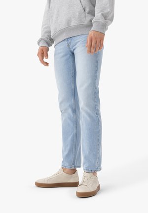 Persoon draagt lichtblauwe recht pijp jeans, grijze sweater en beige sneakers met bruine zolen, staand op een witte achtergrond.