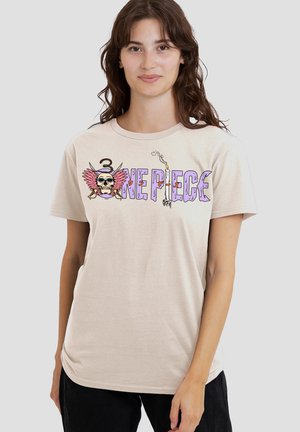 Woman wearing beige T-shirt with "One Piece" text and skull with pink wings graphic, standing against a plain light background.
