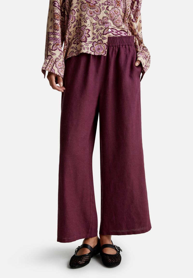 Person, die weite, lila Hosen, eine cremefarbene und lila blumenmusterierte Bluse mit langen Ärmeln und schwarze Mary Jane-Schuhe trägt.