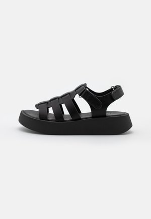 Black platform sandals with a rubber sole, featuring crossed straps and a back heel support. Smooth texture with minimalistic design.