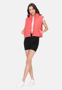 Gilet cropped in corallo brillante con colletto alto, zip frontale e due tasche, abbinato a pantaloni neri e sneakers bianche.