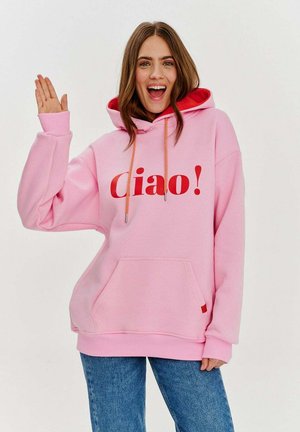 Růžová mikina s červeným nápisem "Ciao!", klokání kapsou a šňůrkovou kapucí. Kombinována s modrými džínami, což ukazuje uvolněný střih a měkkou tkaninu.