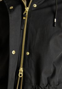 Schwarze Stoffjacke mit goldenen Beschlägen, einem Reißverschluss auf der Vorderseite und Druckknöpfen. Strukturiertes Material mit glatter Oberfläche.
