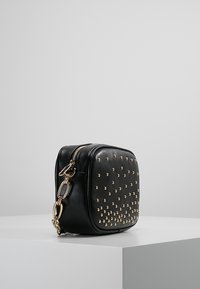 Černá kožená crossbody taška se zlatými hroty, čtvercového tvaru, s horním zipem a zvýrazněným zlatým řetízkovým popruhem.