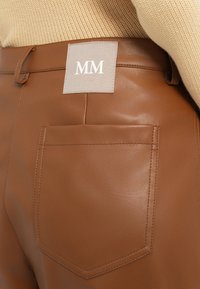 Pantaloni in finta pelle marrone con un'etichetta quadrata di tessuto con "MM" e una tasca posteriore con dettagli cuciti. Texture liscia e forma sagomata.