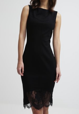 Robe noire sans manches longueur genou avec silhouette ajustée et détails de dentelle transparente à l'ourlet, portée par une femme sur fond gris.