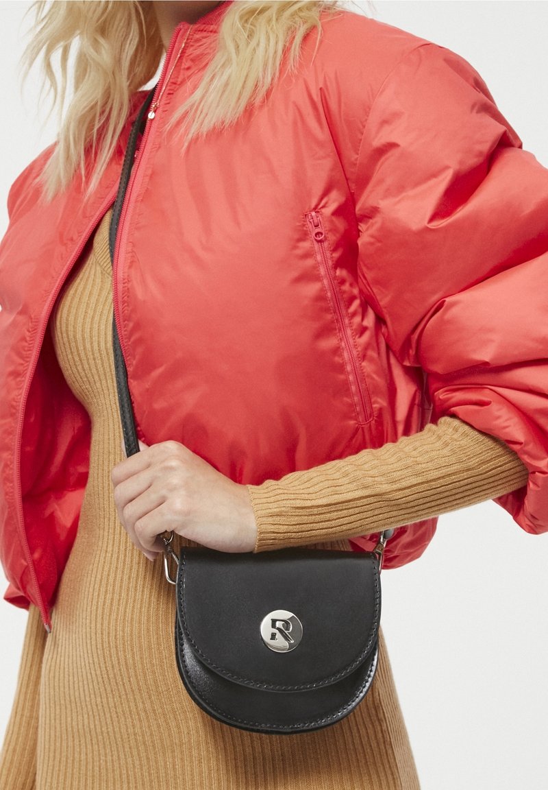 Sac bandoulière en cuir noir de forme ronde avec un logo accent argenté, tenu contre un modèle portant une veste matelassée rouge vif et une robe côtelée beige.
