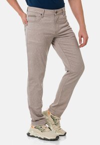 Beige gemusterte Jeans mit slim fit, fünf Taschen, metallischer Knopfverschluss und subtiler Textur, kombiniert mit hellen Sneakers.