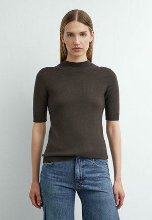 Frau mit glattem hellbraunem Haar, die einen dunkelbraunen Kurzarm-Pullover und blaue Jeans trägt und vor einem schlichten hellen Hintergrund steht.