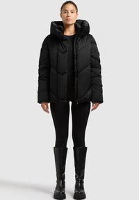 Schwarze Steppjacke mit Zickzackmuster, übergroßer Kapuze und Reißverschluss. Kombiniert mit schwarzen Leggings und kniehohen schwarzen Stiefeln.