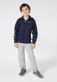 Casaco com fecho de correr azul-marinho com logótipo no peito e na manga, combinado com calças de fato de treino cinza claro. Modelo usa ténis desportivos verdes com detalhes laranja.