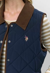 Prešívaná námornícka vesta s hnedým kordovým golierom, zapínaním na cvoky a vyšitým logom. Obsahuje diamantový vzor a bezrukávový dizajn.
