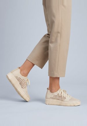 Persona con pantalones beige y zapatillas tejidas color crema con suelas gruesas y texturizadas, un pie plano y el otro levantado atrás.