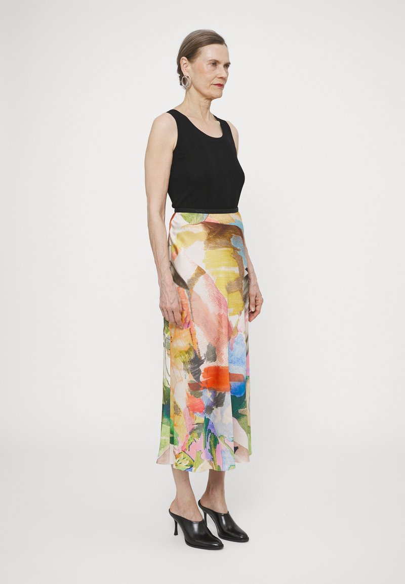 Un haut sans manches noir associé à une jupe longue en soie multicolore aux motifs abstraits. Des mules noires à talons complètent le look.