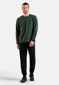 Dunkelgrünes langärmliges, strukturiertes Hemd kombiniert mit schwarzen, taillierten Hosen, die ein enganliegendes Design und elastische Bündchen haben. Schwarze Schuhe runden den Look ab.