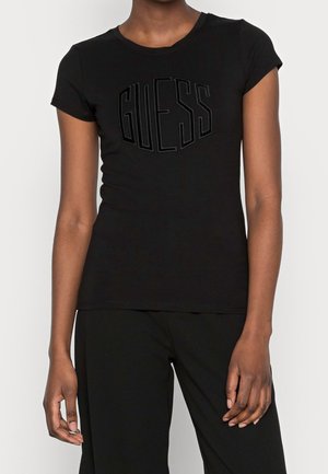 Person trägt ein eng anliegendes schwarzes T-Shirt mit eingeprägtem "GUESS"-Logo und schwarze Hosen, gezeigt von Hals bis Hüfte vor weißem Hintergrund.