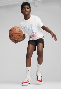 T-shirt bianca con design grafico colorato, pantaloni corti sportivi neri, calze bianche e scarpe sportive con dettagli rossi. Tiene in mano un pallone da basket.