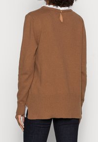 Brązowy sweter z dzianiny z prążkowanym dołem i długimi rękawami. Detal z kluczykiem z tyłu zapinany na złoty guzik. Miękka textura i luźny krój.