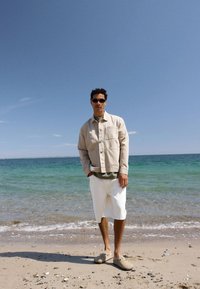 Άνδρας με γυαλιά ηλίου, μπεζ μπουφάν, λευκό σορτς και παντόφλες στέκεται σε αμμώδη παραλία με ήρεμη θάλασσα και καθαρό ουρανό στο φόντο.