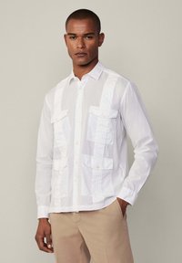 Camisa blanca de manga larga con dos bolsillos frontales, paneles texturizados, cuello abotonado y corte estándar. Combinada con pantalones beige.