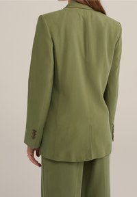 Zelený přizpůsobený blazer se strukturovanými rameny, hladkým povrchem a třemi knoflíky na každém rukávu. Pohled zezadu, který ukazuje čisté linie.