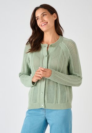 Donna sorridente, indossa un cardigan verde chiaro a maglia aperta con bottoni e jeans azzurri, con le mani leggermente intrecciate davanti.