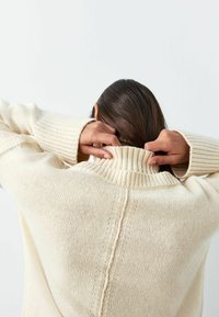 Kremfarvet sweater med høj ribbet krave og tekstureret strik. Har faldende skuldre og en central søm, der løber ned ad ryggen.