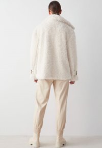 Abrigo exterior blanco, esponjoso y texturizado, con un cuello ancho y mangas largas, combinado con pantalones beige ajustados que tienen puños elásticos.