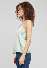 Camiseta de satén verde claro con tirantes finos, escote en pico y un ajuste relajado, combinada con jeans de mezclilla azules. Textura suave en toda la prenda.