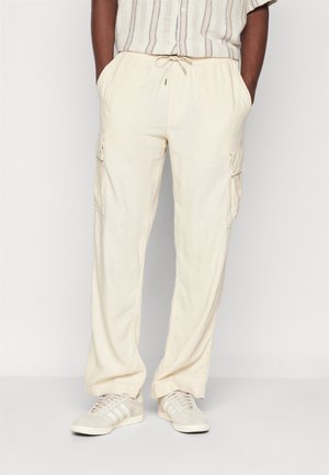 Mann trägt off-white Cargo-Hose mit Seitentaschen, ein gestreiftes Kurzarmhemd und beige Sneaker, steht mit den Händen in den Taschen.