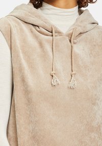 Närbild av en beige kordfluwel utan ärmar med dragsko som bärs över en ljus polotröja, som visar tygtextur och knutdetaljer.